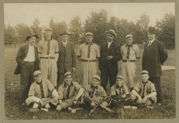 A sepia image of seven men standing and five sitting in front of them posing for a team photo. They are in a park and eight of the men are wearing a uniform.