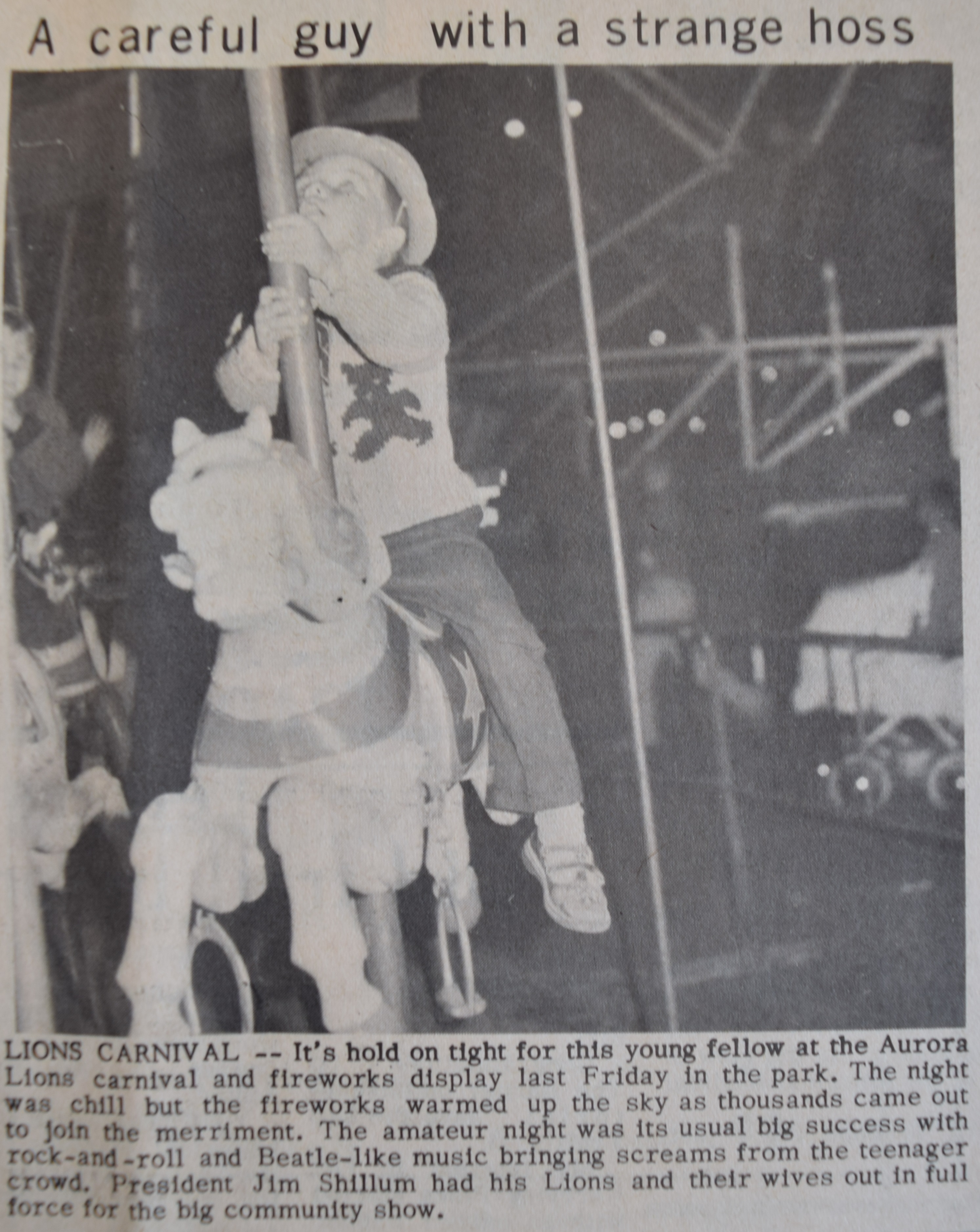 A black and white clipping from a newspaper depicting a photograph of a small boy on a merry-go-round horse; the young boy wears a hat and is looking up; a baby carriage, people and a ferris wheel are visible in the background.