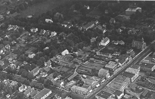 A black and white aerial image showing six blocks of a town. A variety of public and residential buildings can be seen as well as a park in the top left hand corner.
