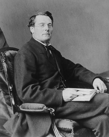 A black and white photograph of a man seated on an arm chair and holding an open book in his lap; he wears glasses and a suit