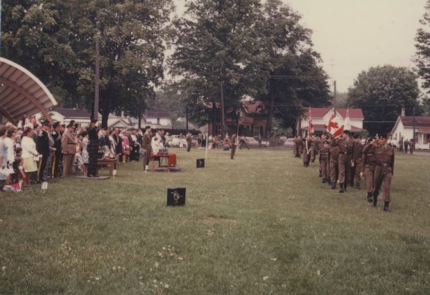 A colour image with a row of cadets marching in a park. They are close to the right hand side of the photo and are marching in the direction of the person taking the photo. On the left hand side of the image there is a crowd of onlookers who are watching the cadets and the roof of a bandshell is visible behind them. In the background some houses are visible as well as mature trees.