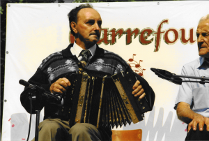 Colour photograph of André Labonté, performing on an outdoor stage. He is sitting on a chair, wearing a cardigan and a tie, as he plays the accordion. Behind him, a few letters of the word “Carrefour” are visible.