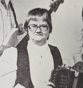 Photograph showing Francine Desjardins seated with an accordion on her lap, surrounded by her band. A musician with a bass guitar can be seen standing behind her.