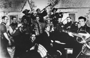The musicians are dressed in traditional garb, some with scarves and others with hats. Behind them is a false backdrop with images of houses.