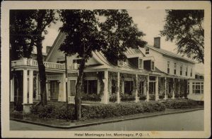 Photograph of a black and white postcard showing the Montmagny Inn at the intersection of two streets. The one-and-a-half storey building has a curved roof featuring three dormer windows. It is surrounded by a covered veranda. A two-storey addition with a gable roof can be seen on the right.