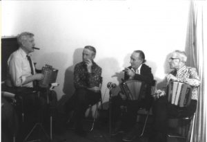Black and white photograph of Philippe Bruneau sitting on a piano bench, accordion in hand. Alfred Couillard, Lorenzo Picard and Joseph Messervier are watching him play. Several microphones have been placed in front of them.