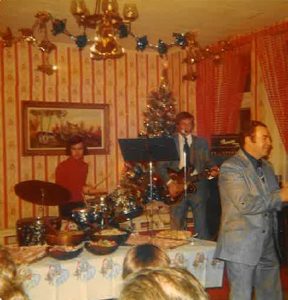 Photograph showing two musicians, in the background, playing at a house party hosted by the Vézina family. In the foreground are the heads of two children who have their eyes riveted on the musicians. They are separated from the musicians by a table on which food has been laid out. On the far right, a man is dancing.
