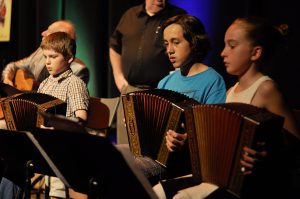 Photograph of three young accordionists performing on stage. They are sitting behind music stands, which hold their sheet music. Their teacher is at the back, watching them.