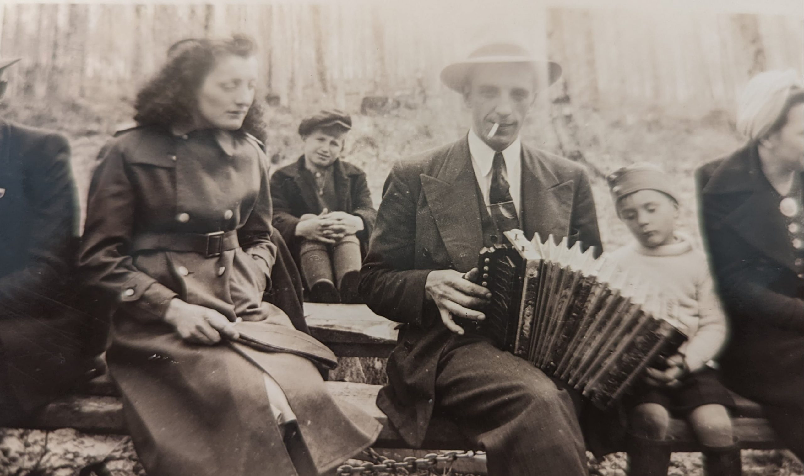 Black and white photograph of accordionist Fernand Fraser playing an accordion on his lap, during a sugaring-off party. A woman is seated next to him, and a child is in the background.