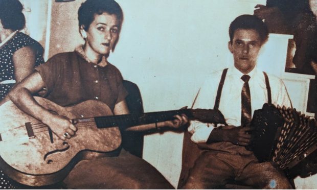 Sepia photograph showing the husband and wife duo of Yvette Coulombe (guitar) and Ulric Coulombe (accordion). Seated on chairs, the musicians are playing at a family dance party.