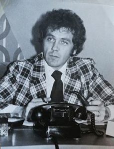 Black and white photograph of Gilles Vallière, pipe and pencil in hand, sitting behind a desk. A telephone can be seen on the desk.