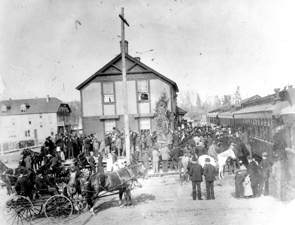 Black and white photo of a large crowd of well-dressed people at two-storey train station with a stationary train on the right. In the crowd, there are horses and carriages.