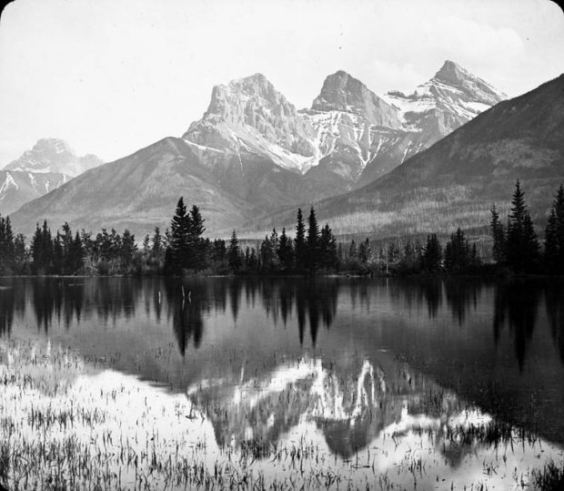 Black and white photo of three snow-covered dramatic peaks with a mirrored lake in front.
