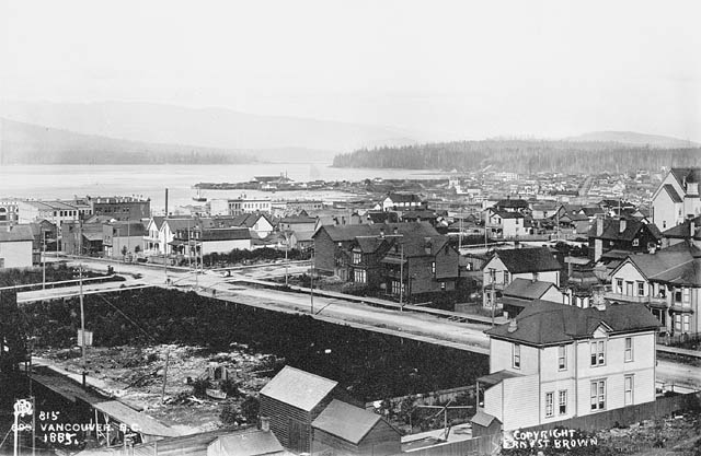 Black and white photo of a young coastal city with the ocean and mountains in the background. Early one and two storey buildings, a large empty lot and roads are in the foreground.