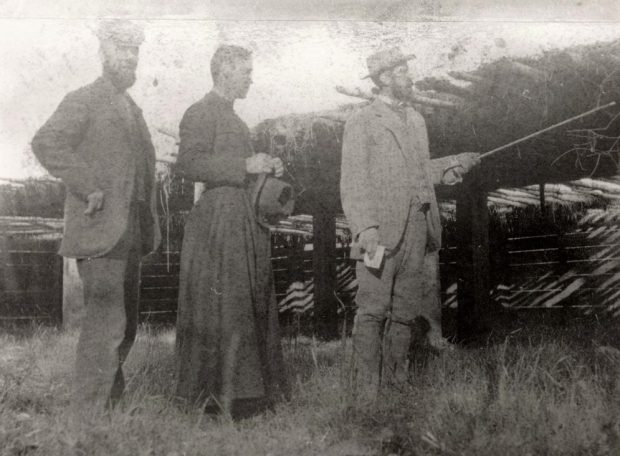 Black and white photo of three men outside. Two are in suits and the man to the right is pointing a walking stick. The man in the centre is wearing a cassock and holding a hat.