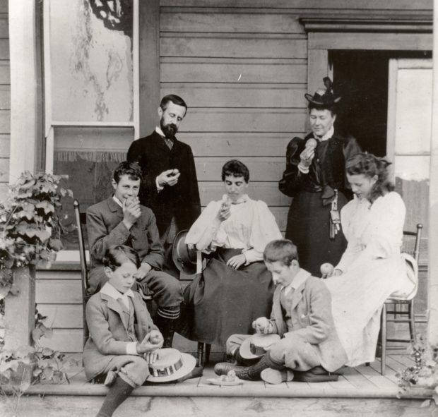 Black and white photo of a family sitting on a verandah. One man, two women, one girl, and three boys each looking at an apple in their hand.