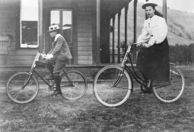 Black and white photo of a girl and a young boy riding bicycles near a house.