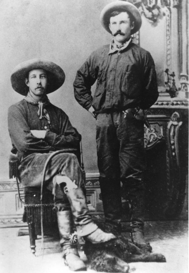 Black and white photo of two men in cowboy clothes and hats. One seated, one standing.
