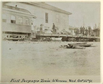 Black and white train photo of part of a passenger car and flatbed car with people standing on It, with a building behind.