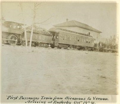 Black and white photo of a train, engine with passenger car and a building behind.