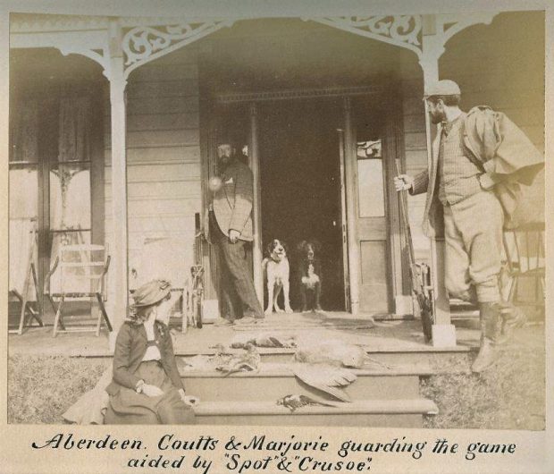 Black and white photo of two men, a girl, two dogs and dead birds on a porch. The man on the right is holding a rifle.
