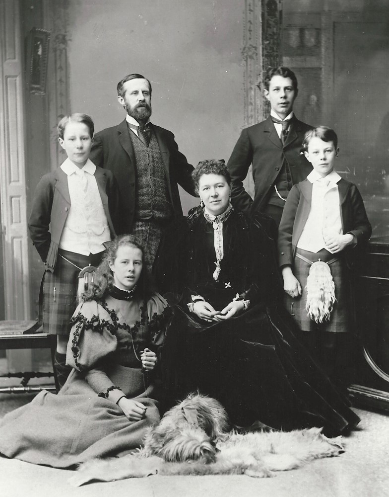 Black and white formal photo of a Victorian era family, a man, a woman, four children and a dog. Two of the boys are in kilts.