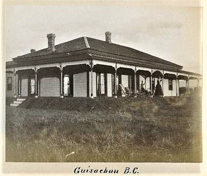 Photo en noir et blanc représentant la façade et le côté d’une maison à étage entourée d’une véranda. Une femme, debout sur la véranda, fait un signe de la main.