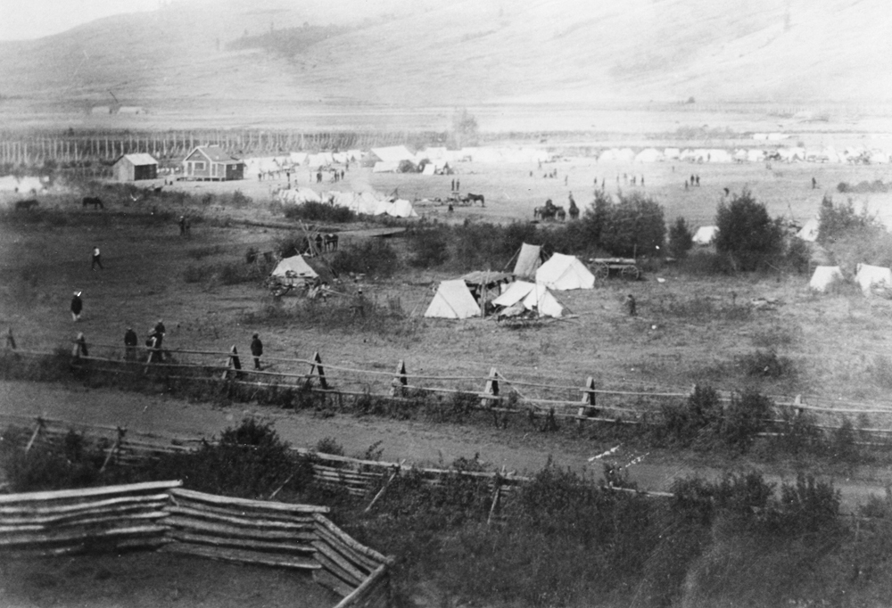 Photo en noir et blanc montrant une clôture, des tentes blanches, une maison, plusieurs petits édifices, des personnes et des chevaux dans une vallée.