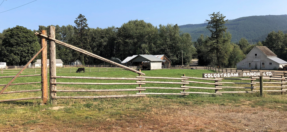 Photo en couleur représentant la clôture d’une ferme avec une pancarte, des champs avec une vache et quatre granges.