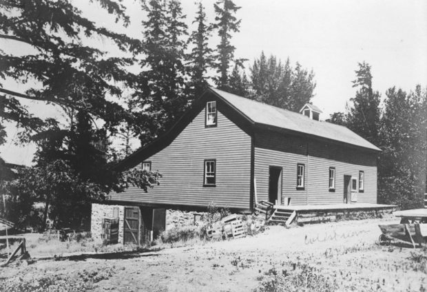 Black and white photo of a one and a half storey building set into a hill. The lower back of the building has a stone foundation and barn doors.