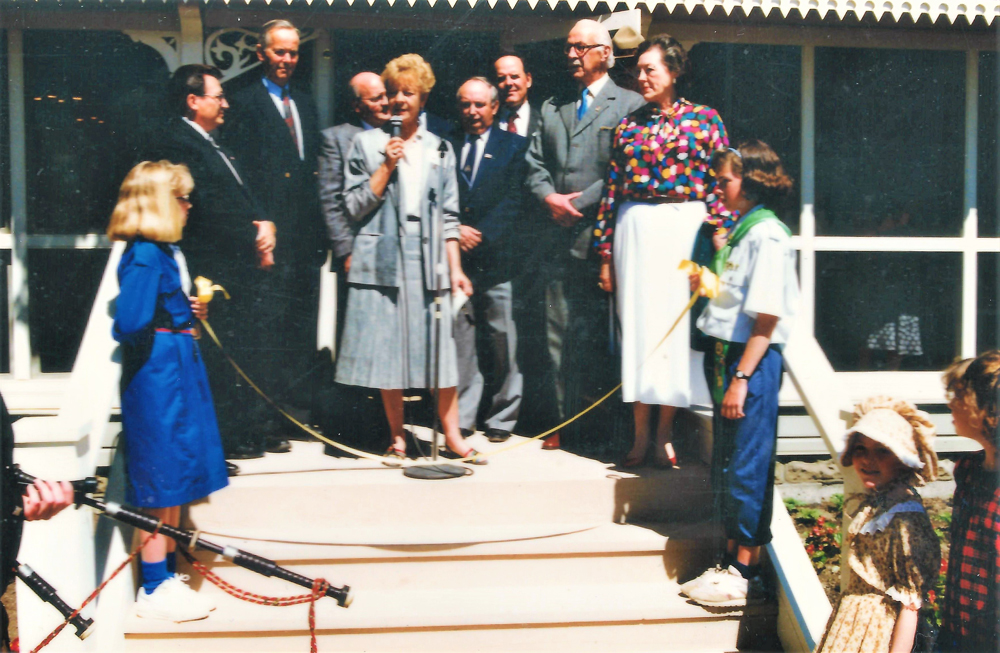 Colour photo, a group of people standing on the verandah. A woman in the middle is speaking into a microphone.