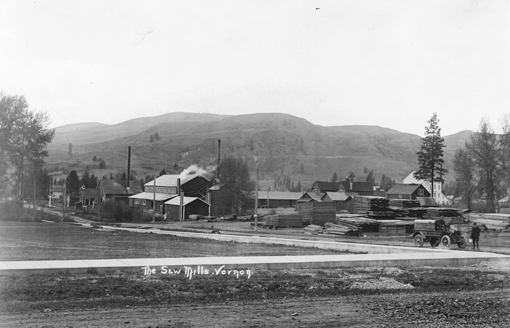 Black and white photo of three small wooden industrial buildings next to a road, with hills behind.