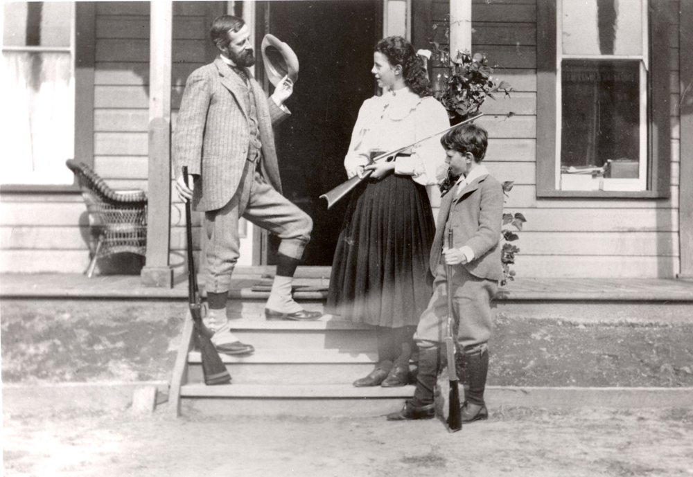 Photo en noir et blanc montrant un homme, une fille et un jeune garçon debout sur les marches d’une maison tenant des fusils de façon décontractée.