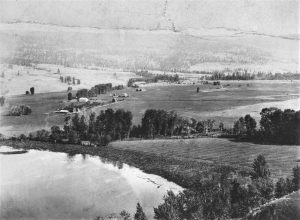 Photo en noir et blanc montrant les bâtiments d’une ferme et des arbres dans le lointain, et un lac dans l’angle inférieur gauche.