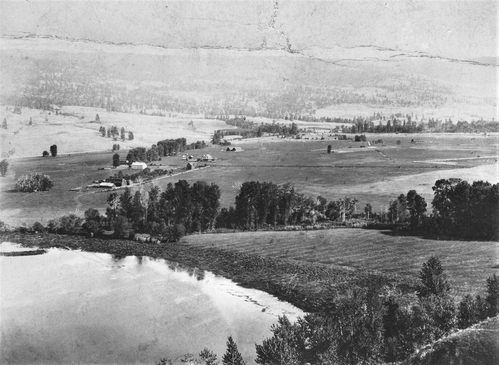 Black and white photo of distant farm buildings and some trees with a lake in the lower-left corner.