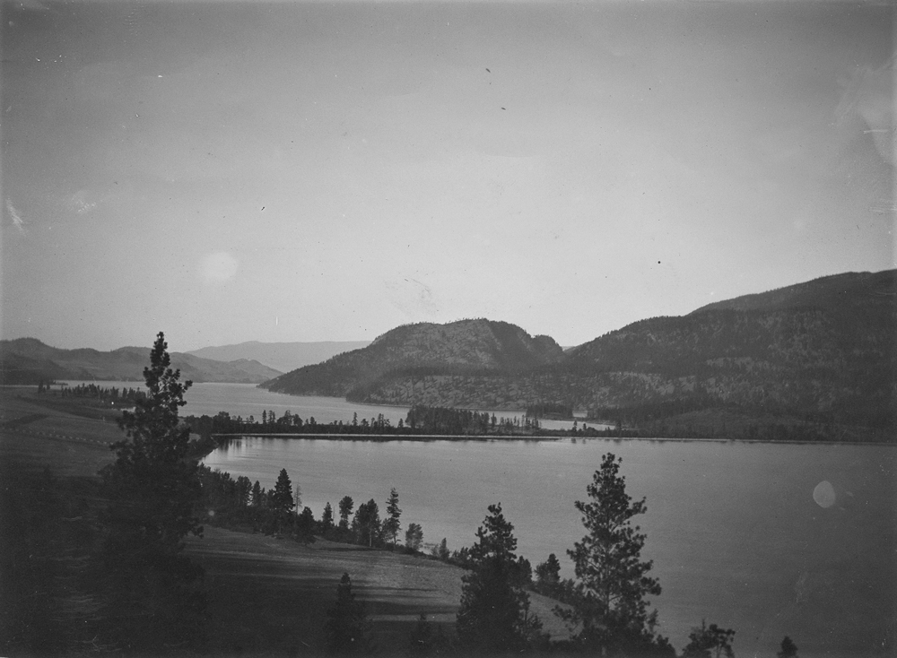 Black and white photo of view of two lakes with a narrow isthmus with low hills in the distance.