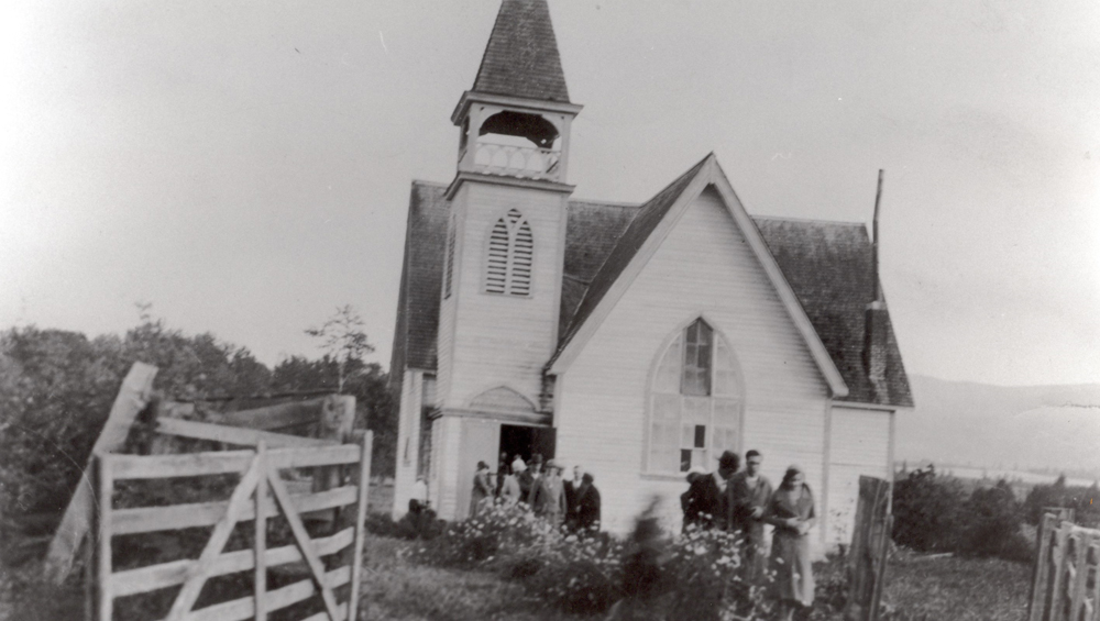 Black and white photo of a group of men and women leaving a small church.