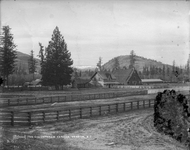 Black and white photo of farm fencing in front of barns and trees with distant hills.