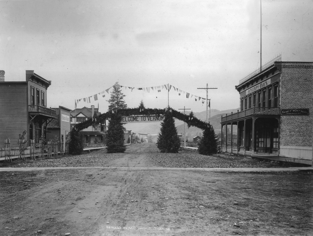 Black and white photo of a gravel road with commercial buildings on each side. Crossing the road is an evergreen Welcome sign.