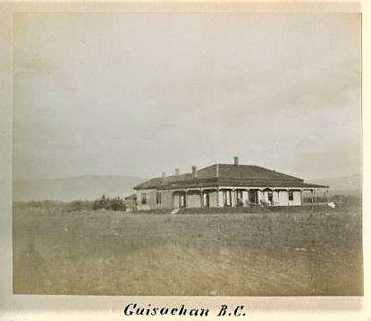 Black and white photo of the front and side of a one-storey house with a wrap-around verandah with a field in front.