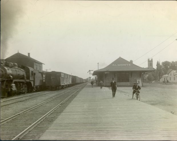 Photograph of Almonte train station with a train on the far track, men standing on the platform, and a man riding a bike next to the platform, c.1930s