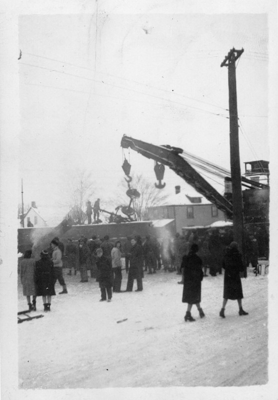 Photograph of the wreckage from the train wreck in Almonte with a crane in the background being used for the clean-up, and people walking through town, 1942