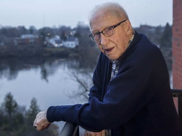 Colour photograph of Douglas Snair in his home, 2017. He is standing on a balcony with housing and a body of water in the background.