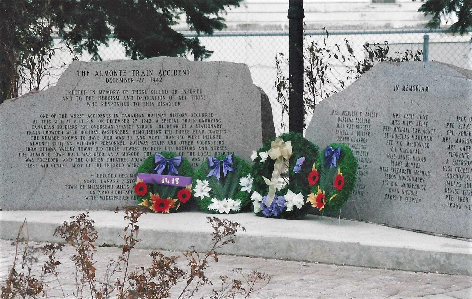 Photographie des deux pierres de granit du monument commémoratif de l’accident ferroviaire d’Almonte, avec quatre couronnes de fleurs déposées au centre, 2000