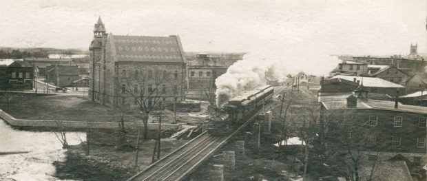Photograph of a train travelling on a track with Almonte town hall in the background, 1950s