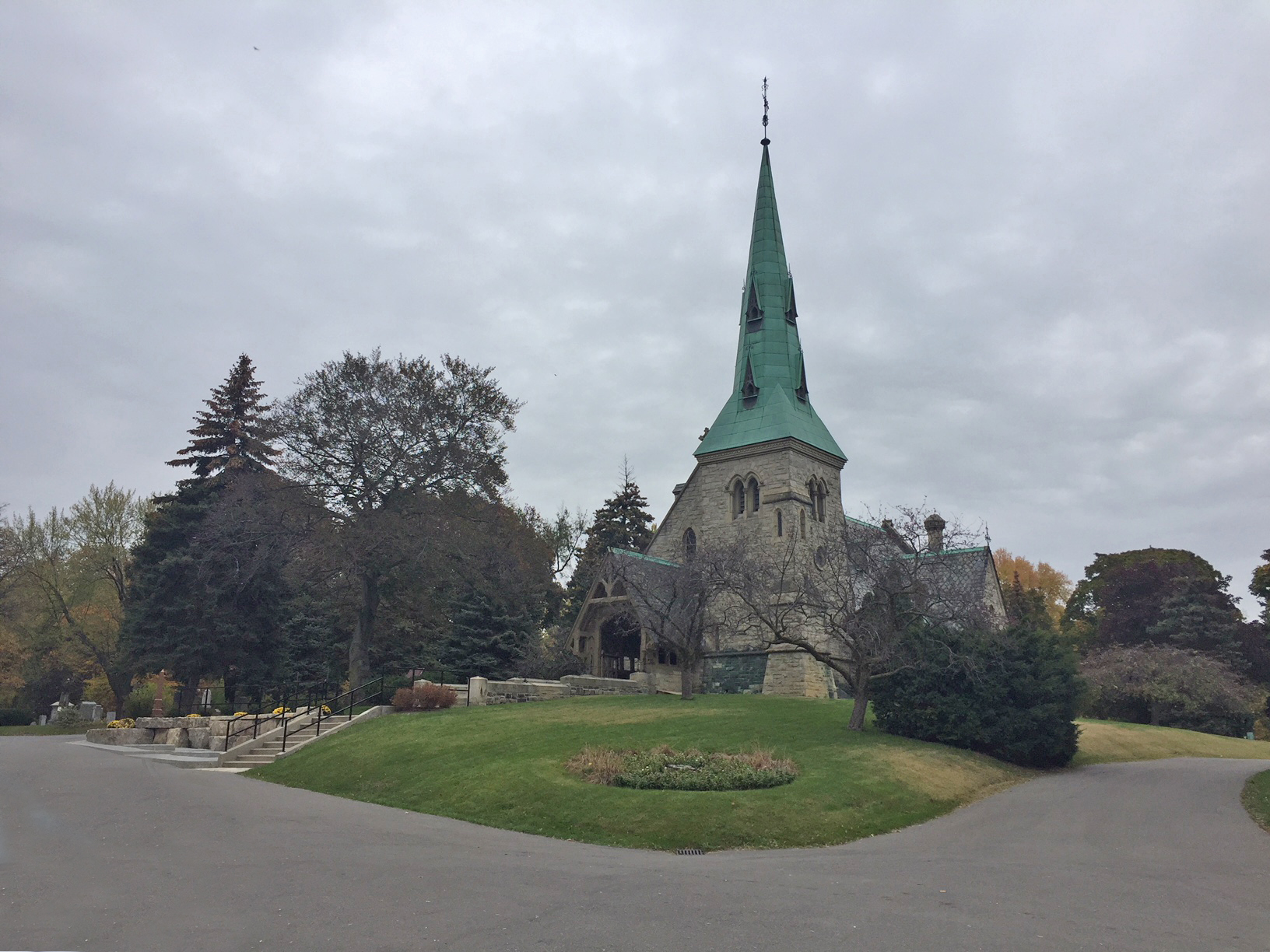 A photograph of St. James 'the Less' Anglican Church in Toronto, Ontario. The stone church has a large bronze steeple that has aged green.