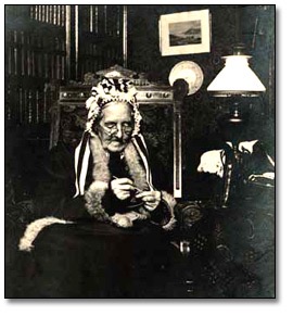Black and White photograph of Anne Langton doing sewing. Miss Langton is seated, with glasses on. She is wearing a black dress, with a shawl around her shoulders and over her head.