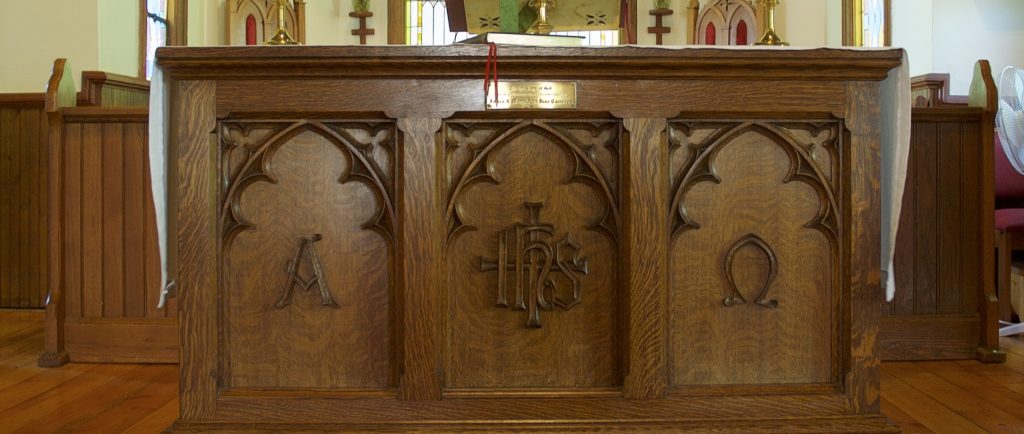 An oak communion table or altar. Left panel has A for alpha.  The middle panel has coat of arms for Church of England. Right panel has upside down U, symbol for Omega.