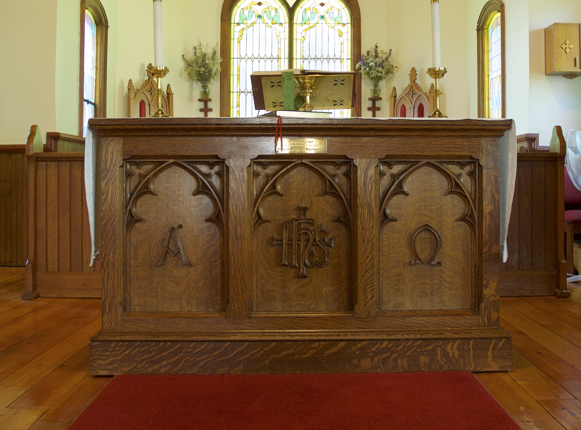 An oak communion table or altar. Left panel has A for alpha.  The middle panel has coat of arms for Church of England. Right panel has upside down U, symbol for Omega.