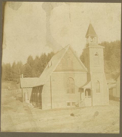 Black and white, and very yellowed, picture of the present day St. James the Apostle. Picture was taken in 1902 at opening. Picture shows the church, the portico entrance and steeple. In the background is the forested hill of the previous churches.
